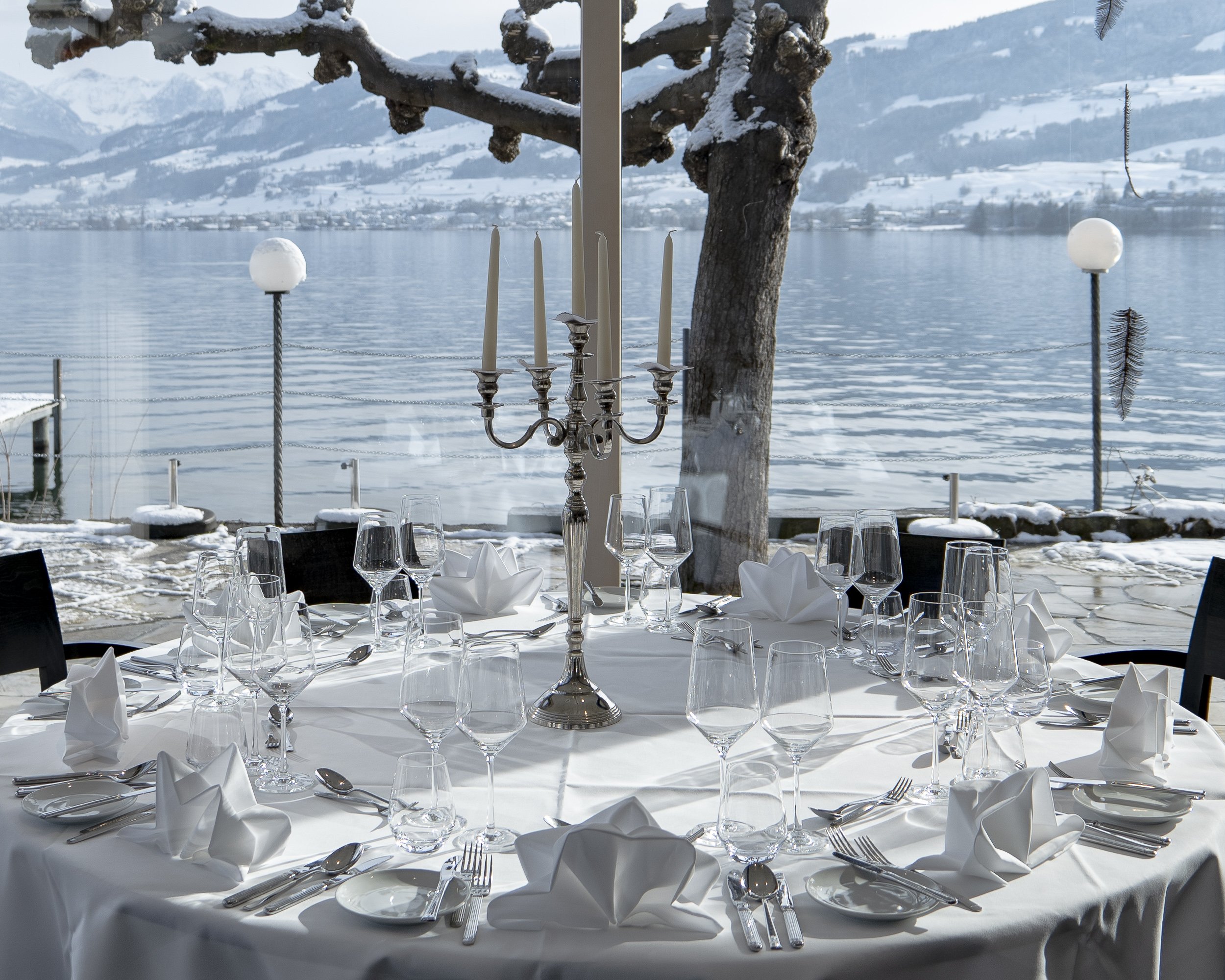 Ein weiss gedeckter Tisch im Seepavillon. Festliches Ambiente mit grossem Kerzenständer. Hinter den grossen Fenstern liegt der winterliche See.